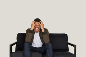 Mad looking young man sitting on the sofa and touching his head with hands