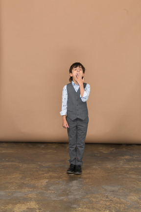 Vista frontal de un niño en traje bostezando y tapándose la boca con la mano