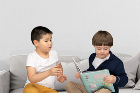 Two young boys sitting on a couch reading a book