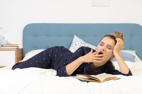 Front view of a yawning young female reading book in bed