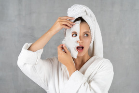 Mujer en bata de baño sosteniendo una máscara de hoja sobre la mitad de la cara