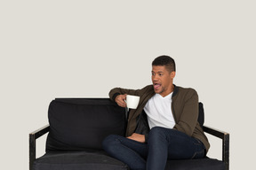Front view of a funny young man sitting on a sofa with a cup of coffee