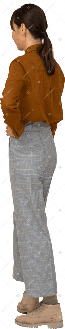 Vista posteriore di tre quarti di una giovane donna asiatica imbronciata in calzoni e camicetta che mette le mani sui fianchi
