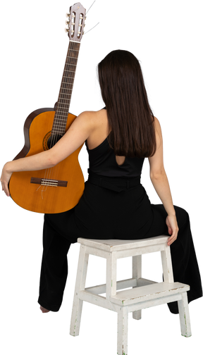 Вид сзади молодой женщины в черном костюме, держащей гитару и сидящей на стуле