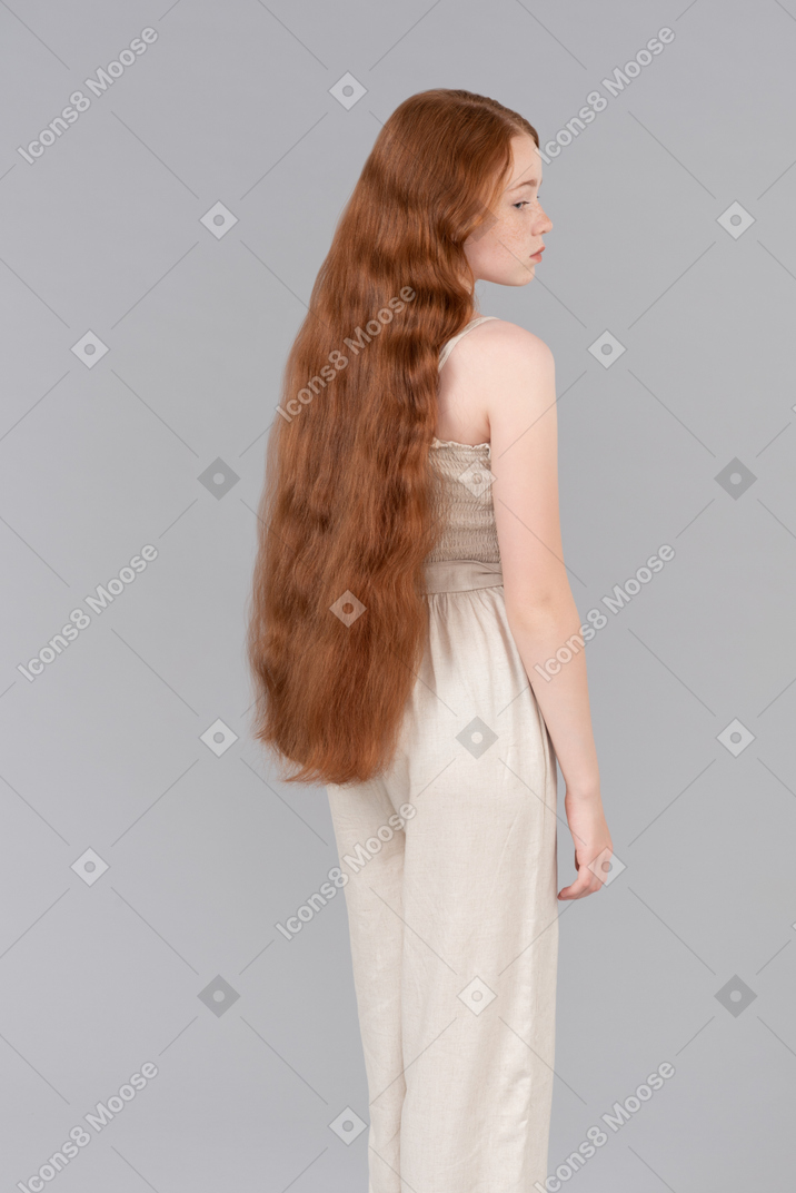 Teenage red haired girl in beige overalls standing half sideways back to camera