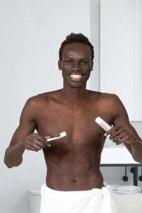A man with a towel around his waist holding toothbrushe and toothpaste