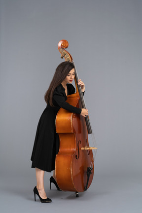 Vista lateral de uma jovem musicista séria de vestido preto tocando seu contrabaixo