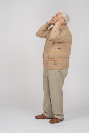 Side view of an old man in casual clothes looking up and covering ears with hands