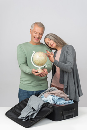 Dreamy middle aged couple holding globus standing next to open suitcase
