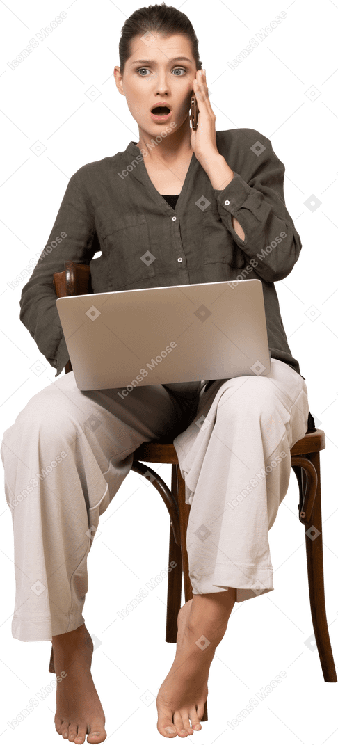 Vista frontal de uma jovem chocada sentada em uma cadeira com um laptop e celular