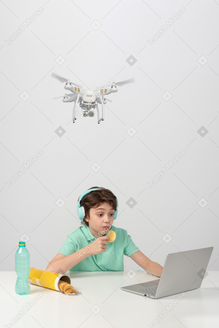 A boy in earphones is so engrossed  in his laptop that he doesn't notice a hovering drone