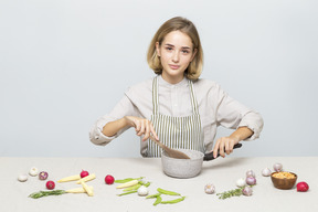 Chica en delantal sosteniendo una cuchara de madera y sentada en la mesa con pan y verduras en ella