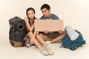 Young interracial couple of hitchhikers holding paper card and showing hitchhiking gestures