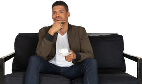 Vista frontale di un giovane uomo pensieroso seduto su un divano con una tazza di caffè