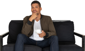 Front view of a young thoughtful man sitting on a sofa with a cup of coffee