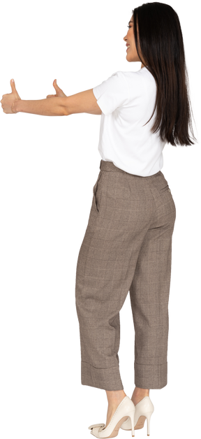 Vista posterior de tres cuartos de una joven sonriente en calzones y camiseta mostrando los pulgares hacia arriba