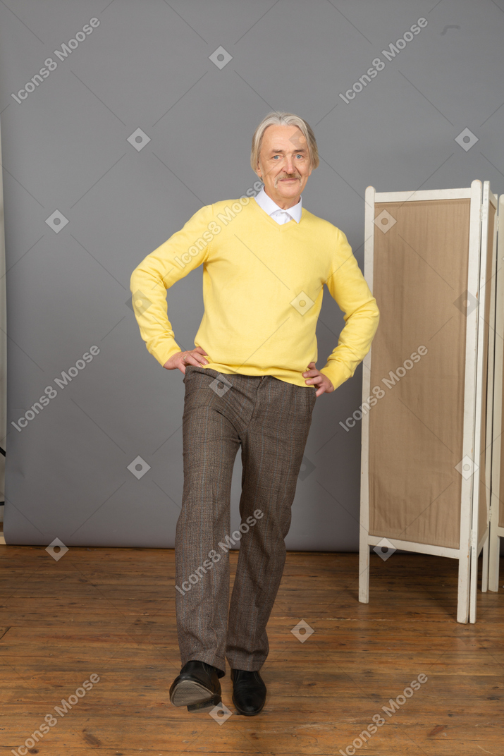 Vista frontal de un anciano sonriente poniendo la mano en las caderas mientras levanta la pierna
