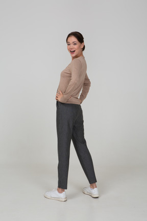 Vista posteriore di tre quarti di una giovane donna sorridente in pullover e pantaloni mettendo le mani sui fianchi