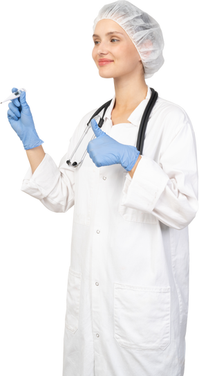 Трехчетвертный вид улыбающейся молодой женщины-врача со стетоскопом, держащей термометр и показывающей большой палец вверх