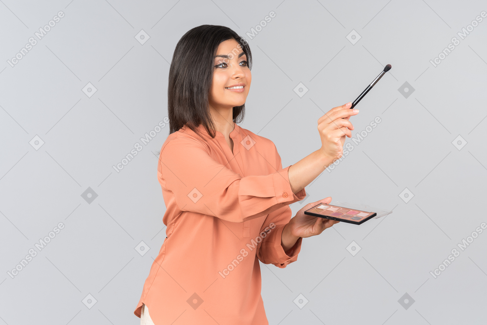 Smiling indian girl standing half sideways and holding eye brush and eyeshadow palette