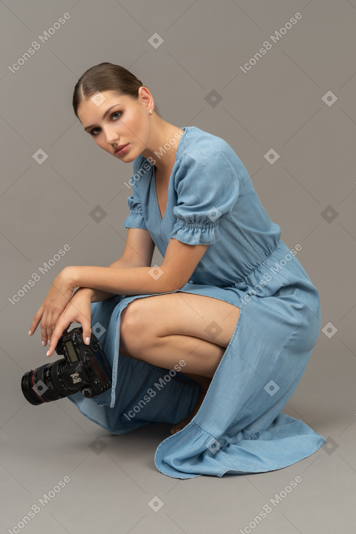 一位身穿蓝色连衣裙的年轻女子坐在地板上，拿着相机的侧视图