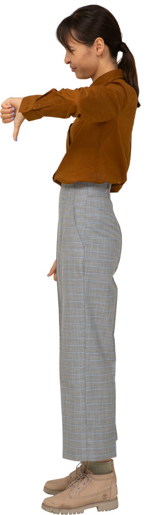 Vista laterale di una giovane donna asiatica in calzoni e camicetta che mostra il pollice verso il basso