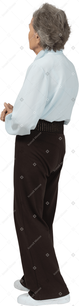 Vista posteriore di una donna anziana seria vestita in camicetta e pantaloni che tengono le mani insieme