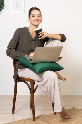 Front view of a smiling young woman sitting on a chair and holding her laptop & coffee cup