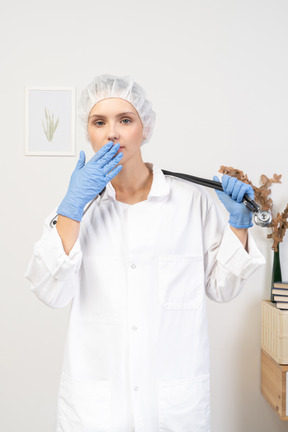 Vista frontal de una joven doctora sosteniendo un estetoscopio y escondiendo la boca