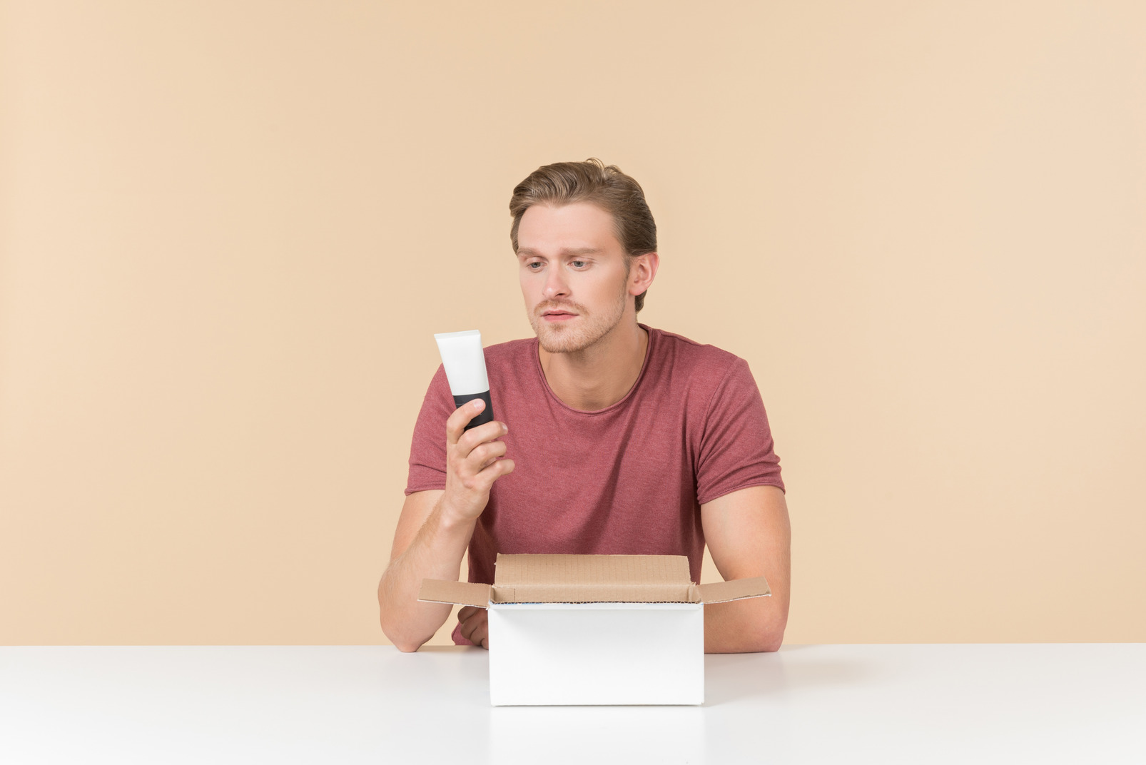 Young guy sitting at the table with box on it and looking at cream tube