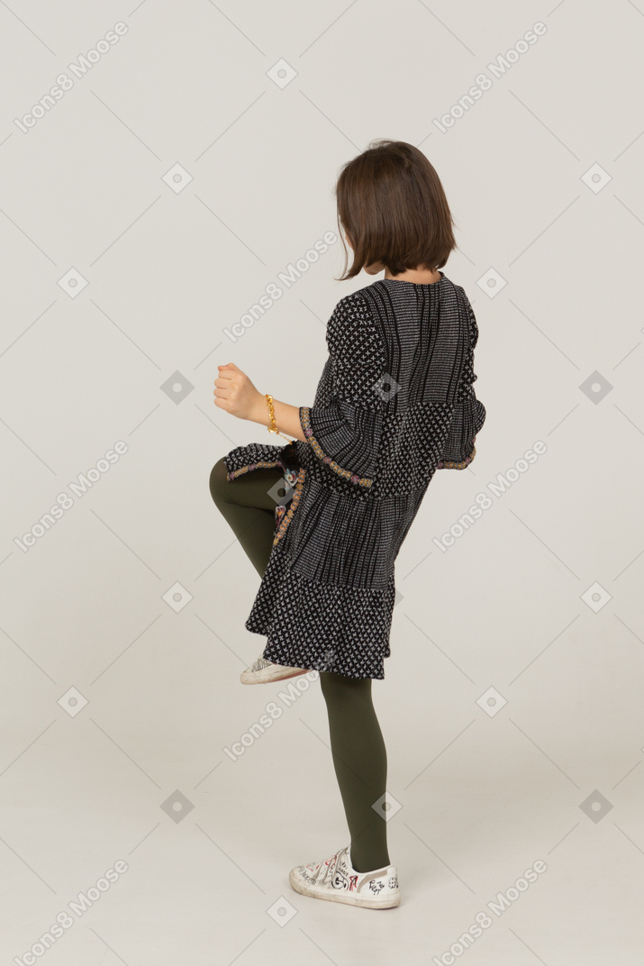 Three-quarter back view of a happy little girl in dress clenching fists and raising leg