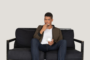 Front view of young man sitting on a sofa and holding cigarette and a cup of coffee