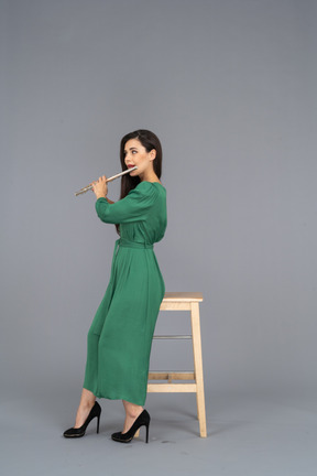 Vista lateral de una joven en vestido verde sentada en una silla mientras toca el clarinete