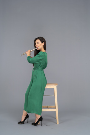 Side view of a young lady in green dress sitting on a chair while playing the clarinet