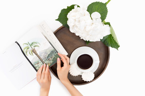 Cup of coffee, meringues, white flower and book on round wooden tray