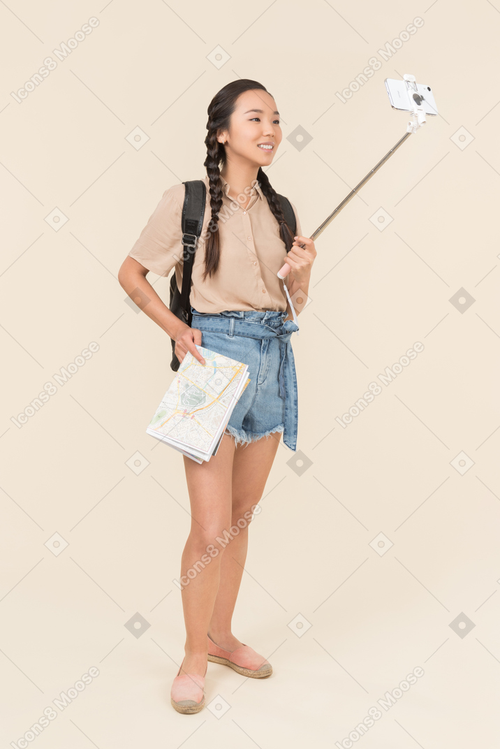 Young female traveller holding city map and making a selfie with smartphone on selfie stick