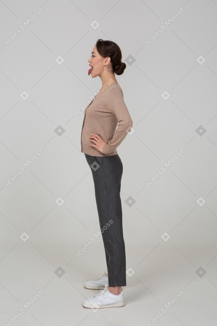 Side view of a young lady in pullover and pants putting hands on hips and showing tongue