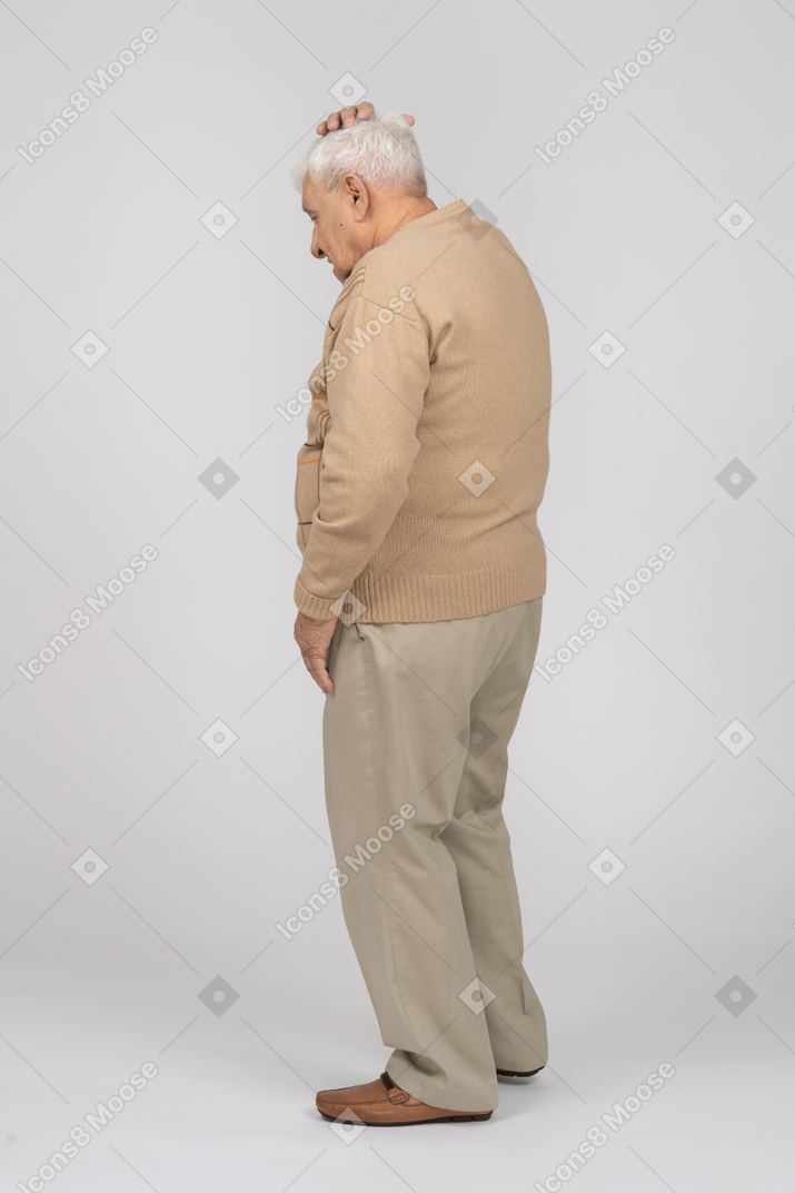 Side view of an old man in casual clothes standing with hand on head and looking down