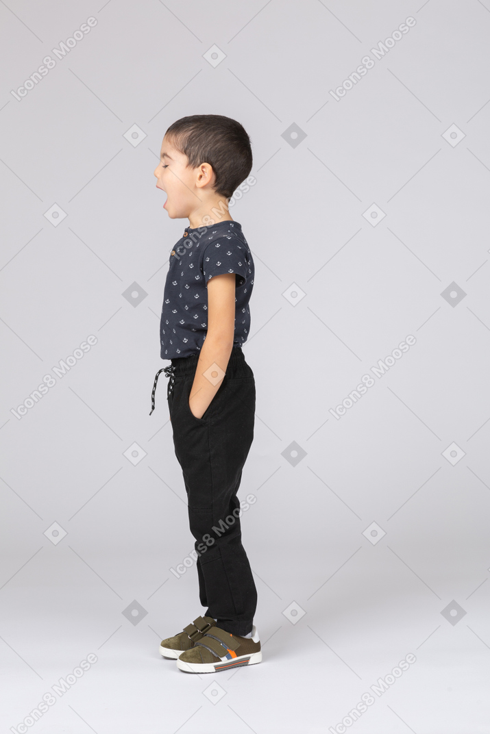 Side view of a sleepy boy standing with hands in pockets and yawning