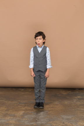 Front view of a cute boy in grey suit standing still and making faces