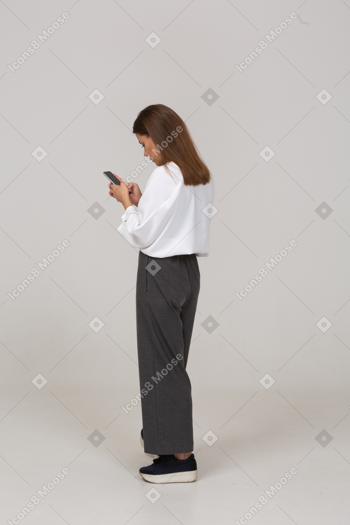 電話でフィードをチェックするオフィス服の若い女性の4分の3の背面図