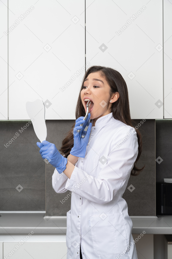 Vista de três quartos de uma médica assustada, olhando-se no espelho e tocando os dentes com um instrumento dentário
