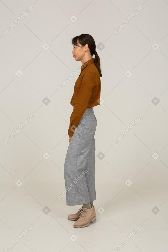 Vista lateral de uma jovem mulher asiática fazendo beicinho, de calça e blusa, parada