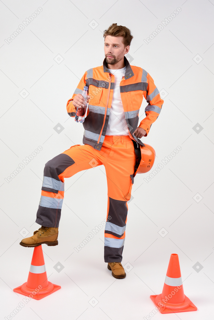 Handsome street worker standing with his leg on cone and drinking water