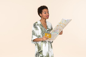 Mujer negra de pelo corto con un vestido blanco y verde, de pie con un pasaporte internacional y un mapa en las manos