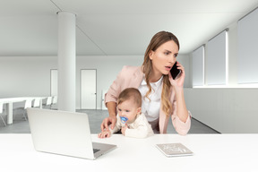 A woman holding a baby in front of a laptop and talking on the phone
