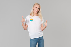 Beau jeune homme aux longs cheveux blonds, debout sur un fond gris, vêtu d'un jean bleu et d'un t-shirt blanc avec écusson lgbt