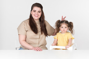 Mãe e filha sentada na mesa e mãe fazendo orelhas de coelho para a filha