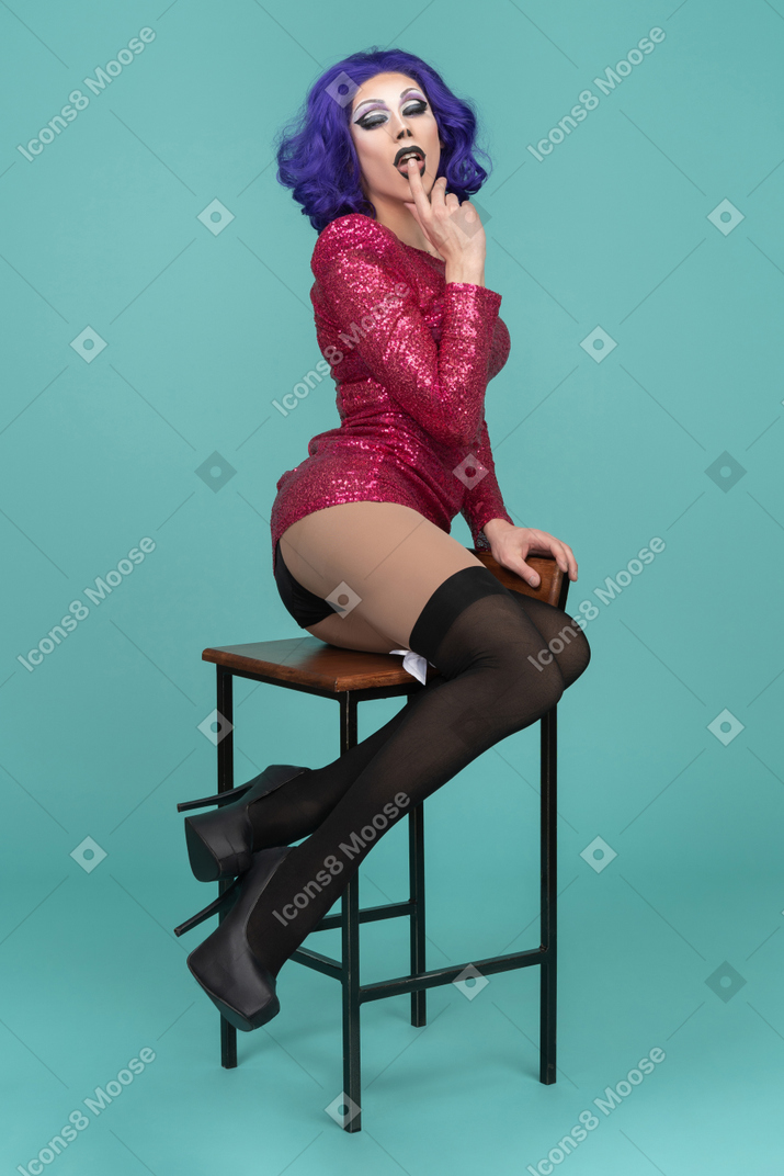 Drag queen pressing finger to lips while sitting on a stool