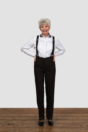 Une vieille femme souriante vêtue de pantalons et de bretelles souriant et mettant les mains dans les hanches